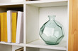Round faceted green glass vase on a square white bookshelf. Glass object to decorate the space. Living room or office interior. Details of modern design.