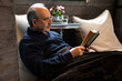 © YKD - Elderly man with a mustache and reading glasses covered with a soft blanket sits on a bean bag and reads a book in cozy interior of wooden house