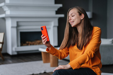 Adorable brunette Spanish woman in orange blouse and black pants sitting at living room holding phone making video call ;ooking at screen. Beautiful housewife making selfie at new home. Happy women.