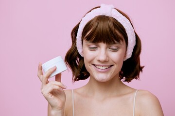 Wall Mural -  aesthetic woman poses covering her eyes with pleasure and holding a white jar of cream in her hand. Horizontal photo on a pink background with an empty space for inserting an advertising layout