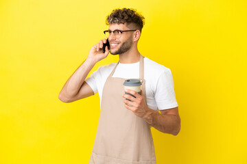 Poster - Restaurant waiter blonde man isolated on yellow background holding coffee to take away and a mobile