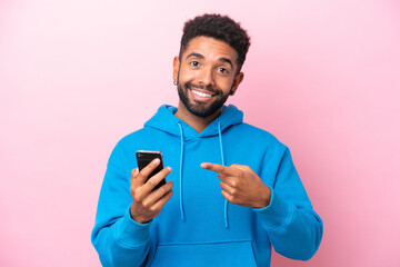 Wall Mural - Young Brazilian man isolated on pink background using mobile phone and pointing it