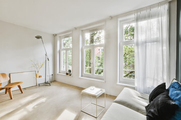 a living room with white walls and wood flooring, two large windows in the wall is covered with sheer curtains