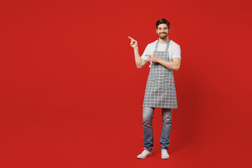 Wall Mural - Full body smiling young male housewife housekeeper chef cook baker man wear grey apron point index finger aside on workspace area mock up isolated on plain red background studio. Cooking food concept.