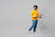 Side profile view full body young smiling happy IT Indian man 20s he wear casual yellow hoody hold use work on laptop pc computer look aside on area isolated on plain grey background studio portrait.