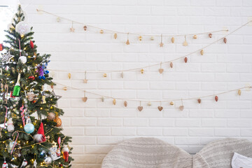 Wall Mural - Christmas tree in white interior of a house with loft-style brick walls with garlands of glass toys on a rope. Glowing fairy lights decoration of the studio room. Cozy home in christmas and new year