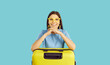 © Studio Romantic - Portrait of happy beautiful young woman traveler with yellow suitcase on light blue background. Stylish casual girl in yellow glasses and earrings leans on suitcase and looks at camera with smile.