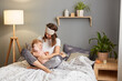 © sementsova321 - Portrait of happy dark haired mother wearing sleeping mask with her baby daughter playing in bedroom while sitting on bed, woman hugging her infant kid with love.