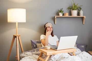 Horizontal shot of sleepy tired woman wearing sleep mask sitting and working on laptop in bed at home, feels sleepless works whole night till morning, yawning, covering mouth with palm.