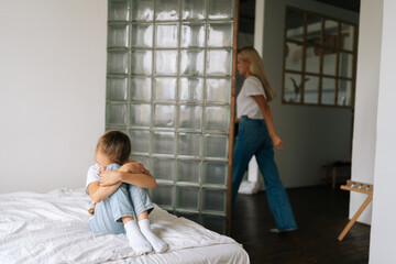 Angry young mother leaving the children room after after quarrel with sad crying little child daughter sitting on bed hugging knee, cover face at home. Concept of family problems, conflict, crisis.