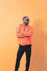 Wall Mural - Handsome African male fashion model wearing sunglasses and seriously posing with crossed arms while looking to the side, he is standing near the orange wall outside. African man smiling over isolated 