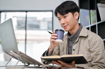 Smart Asian male programmer or web developer sipping coffee and using laptop computer.