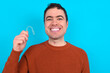© Jihan - Young handsome Caucasian dark haired man standing over blue studio background holding an invisible aligner braces and smiling showing perfect results.