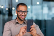 © Liubomir - Close-up of smiling African-American businessman using phone typing messages and browsing internet pages, man in shirt and glasses smiling happily inside office holding smartphone.