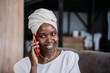 © Iona - Cheerful African young woman in turban sitting on sofa talking by phone toothy smiles looks aside. Happy businesswoman in traditional african clothes discussing with partner using sell phone.