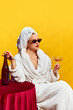 Leinwandbild Motiv Young woman in bathrobe sitting and drinking champagne over yellow background. Feeling relaxed