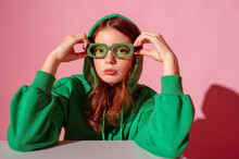 Fashionable Freckled Redhead Girl Wearing Trendy Green Hoodie, Stylish Color Rectangular Glasses, Posing Against Pink Background. Close Up Studio Portrait. Copy, Empty Space For Text
