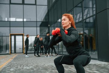 Doing exercises with dumbbells. Group of sportive women is outdoors near black building