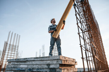 Wall Mural - In grey uniform. Holding wooden plank. Man is working on the construction site at daytime