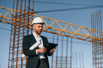 Wall Mural - In white hard hat, with plan and tablet in hands. Businessman in formal clothes is on the construction site at daytime