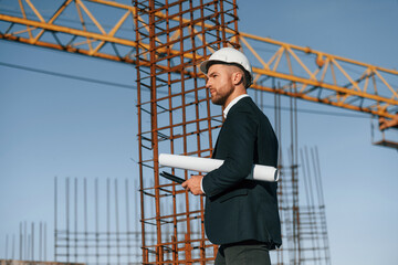 Wall Mural - Side view. Walking and holding paper with plan and digital tablet. Businessman in formal clothes is on the construction site