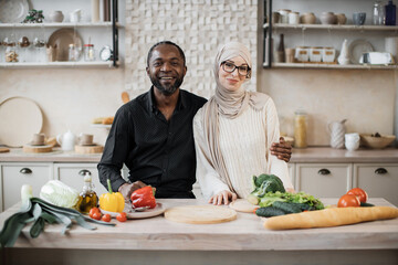 Wall Mural - Young attractive couple in love preparing salad from fresh vegetables. Handsome african man and muslim charming woman cooking dinner together and having fun in a new modern apartment.