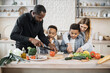 © sofiko14 - Happy multinational parents and two preschool sons preparing tasty food at kitchen. Mother and father teaching lovely kids to cook. Young mom and dad with their children spending time together at home