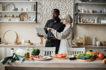 Wall Mural - Happy multiethnic family preparing vegetable salad for breakfast standing near table looking on laptop pc computer at home kitchen. Vegetarian food, healthy eating, people, technology and diet concept