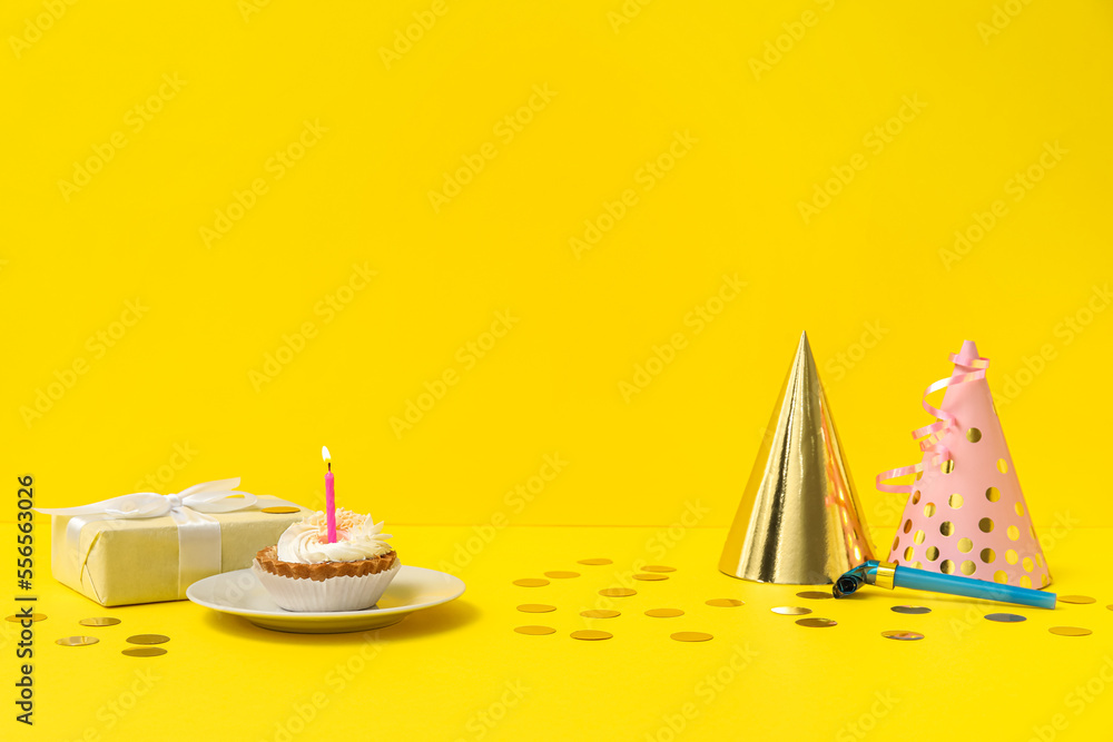Party hats, gift, birthday cake and confetti on yellow background
