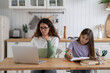 Remote work with homeschooled kids. Focused young woman freelancer working online on laptop while sitting at table in kitchen with school-aged girl daughter doing homework. Freelance and family life