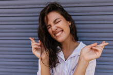 Funny Young Caucasian Girl Stands Outside With Closed Eyes Crossed Fingers For Luck. Model With Brunette Hair Wears White Shirt. Concept Of Good Mood.