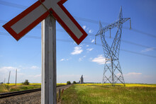 Close-up Of Railway Crossing Sign And Waist-type Transmission Tower In A Canola Field With Train Tracks And Grain Terminal In The Distance; Alberta, Canada