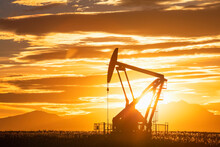 Pumpjack In A Field With A Bright Sun Glowing In The Sky At Sunset And The Distant Silhouetted Rocky Mountains; Alberta, Canada
