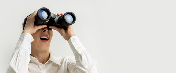 Wall Mural - Surprised young woman in a white shirt looks through binoculars on a white background. Banner