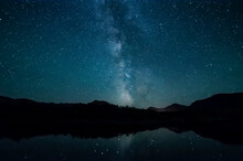 Milky Way And Starry Night Sky Over Silhouetted Mountains Reflected In Water, Waterton Lakes National Park; Alberta, Canada
