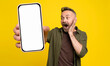 excited middle-age man with hand gesture at face in casual green shirt posing isolated on yellow background looking and showing big smartphone with white mock-up screen holding in hand