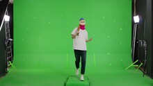 Girl In White T-shirt, Jeans And Sneakers Walking On A Green Screen, Chroma Key. Young Woman In Balaclava With Baseball Bat In Hands. Hooligan Girl In Mask