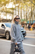 © LIGHTFIELD STUDIOS - trendy woman in grey sweater and sunglasses holding handbag while walking on street of New York city