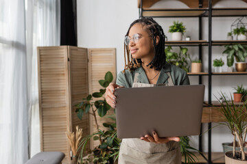 positive african american florist with stylish dreadlocks and eyeglasses holding laptop and looking away in flower shop