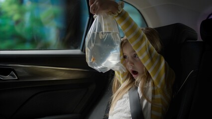 Happy excited Caucasian girl riding on a back seat of a car, bringing home a goldfish in a bag from a pet store