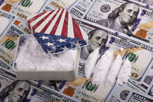 A Small Metal Box With An Image Of The American Flag Is Filled With White Powder, Symbolizing Drugs, Against The Backdrop Of 100 US Dollar Bills.