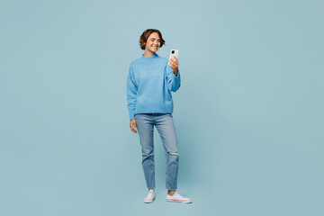 Wall Mural - Full body young smiling happy woman wear knitted sweater look camera hold in hand use mobile cell phone isolated on plain pastel light blue cyan background studio portrait. People lifestyle concept.