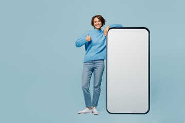 Wall Mural - Full body fun young woman in knitted sweater near big huge blank screen mobile cell phone smartphone with workspace mockup area show thumb up like isolated on plain pastel light blue cyan background