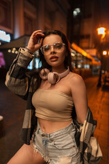 Beautiful sexy woman with trendy glasses and pink headphones in a fashion top with a plaid shirt and jeans ripped shorts walks in the night city with bokeh light
