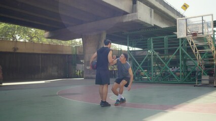 Wall Mural - 4K Asian man basketball player lifting his friend that fallen on outdoors court. Sportsman do sport training streetball at street court under highway in the city. Fair game sport competition concept.