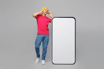 Wall Mural - Full body young man of African American ethnicity in pink t-shirt yellow hat headphones listen music big huge blank screen mobile cell phone with area mockup isolated on plain grey background studio