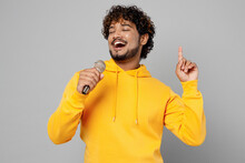 Young Cheerful Indian Man 20s He Wearing Casual Yellow Hoody Sing Song In Microphone On Karaoke Club Stage Point Finger Up Isolated On Plain Grey Background Studio Portrait. People Lifestyle Portrait.