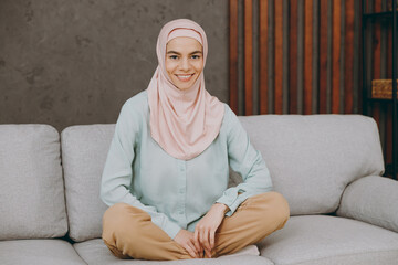 Wall Mural - Full body young muslim woman wear hijab casual clothes sits on sofa couch in lotus pose stay home flat rest relax spend free spare time in living room indoor People uae middle eastern islam concept.