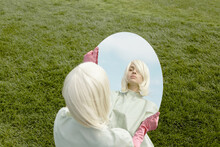 Attractive Woman With Pale Skin And White Blond Hair Wearing Bizarre Costume Looking At Herself In The Mirror On Green Lawn Background