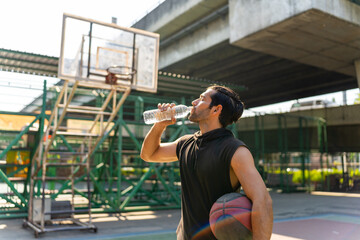 Wall Mural - Caucasian sportsman basketball player drinking and pouring water from a bottle on his face after do sport training and playing streetball on outdoors court under highway in the city in sunny day.